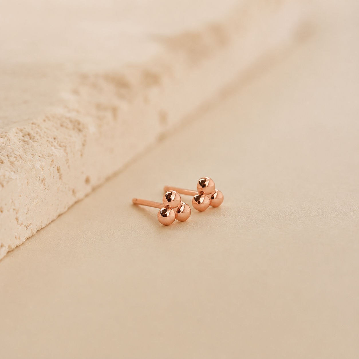 A pair of rose gold stud earrings with three small spherical beads on each earring, displayed on a soft beige textured surface.